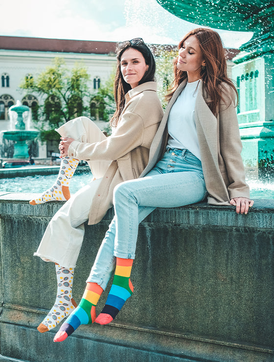 Zwei Frauen sitzen mit bunten Socken an einem Brunnen. Gay Pride Socken mit Regenbogen-Motiv - LGBTQ - Homo - Trans - Queer ✓Hand gekämmte Biobaumwolle ✓Angenehmer Tragekomfort