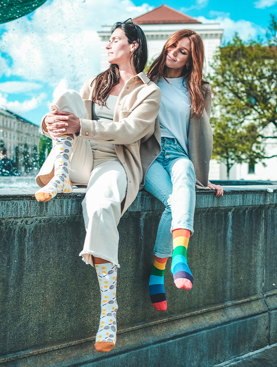 Zwei Frauen sitzen mit bunten Socken an einem Brunnen. Gay Pride Socken mit Regenbogen-Motiv - LGBTQ - Homo - Trans - Queer ✓Hand gekämmte Biobaumwolle ✓Angenehmer Tragekomfort
