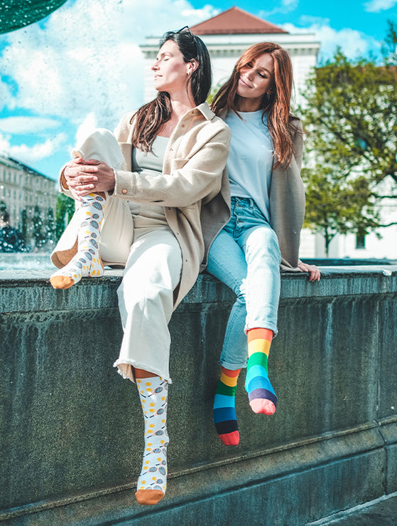 Zwei Frauen sitzen mit bunten Socken an einem Brunnen. Gay Pride Socken mit Regenbogen-Motiv - LGBTQ - Homo - Trans - Queer ✓Hand gekämmte Biobaumwolle ✓Angenehmer Tragekomfort